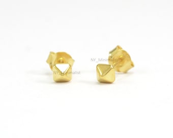 May include: A pair of gold stud earrings with a pyramid-shaped design. The earrings are small and minimalist.