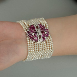 Pode incluir: Um bracelete de pérolas multi-fio com um fecho redondo grande de rubi e diamantes. O fecho é engastado em metal branco e apresenta um design geométrico.