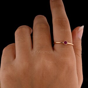 May include: A thin rose gold ring featuring a small, round, deep red ruby gemstone set in a delicate bezel setting. The band is smooth and slender, highlighting the vibrant color of the stone against the metallic finish of the ring's setting.