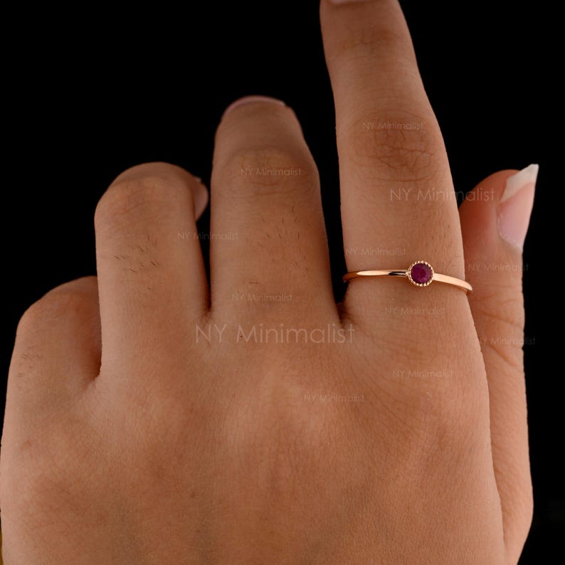 May include: A thin rose gold ring featuring a small, round, deep red ruby gemstone set in a delicate bezel setting. The band is smooth and slender, highlighting the vibrant color of the stone against the metallic finish of the ring's setting.
