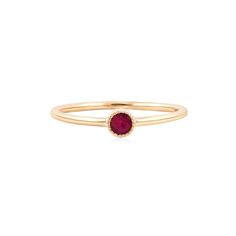 May include: A thin gold band ring featuring a single, round, deep red gemstone set in a delicate, scalloped-edge bezel setting. The ring has a smooth, polished finish and a minimalist design, highlighting the vibrant color of the central stone.