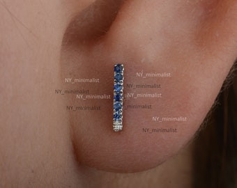 Puede incluir: Un pendiente de barra de plata con una fila de zafiros azules y pequeños diamantes transparentes. El pendiente se muestra en la oreja de una persona.