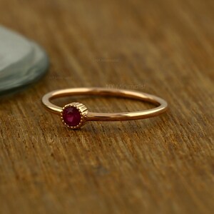 May include: A thin rose gold ring featuring a single, small, round, deep red ruby gemstone set in a bezel setting with a delicate, scalloped edge. The band is smooth and circular, showcasing a polished metallic finish throughout the design.