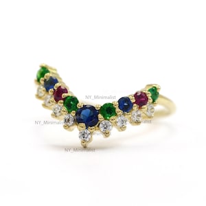 May include: A gold band ring with a V-shaped design featuring a row of small diamonds and colorful gemstones, including blue sapphires, red rubies, and green emeralds.