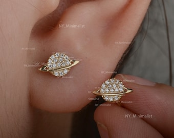 Puede incluir: Un par de pendientes de tono dorado con una pequeña piedra redonda, brillante y transparente, rodeada por un fino anillo dorado, que se asemeja a un planeta con un sistema de anillos.