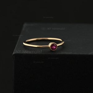 May include: A thin gold ring featuring a single, small, round, faceted pink gemstone set in a beaded bezel. The band is smooth and polished, showcasing a delicate design with a vibrant, circular pink stone as the central focal point.