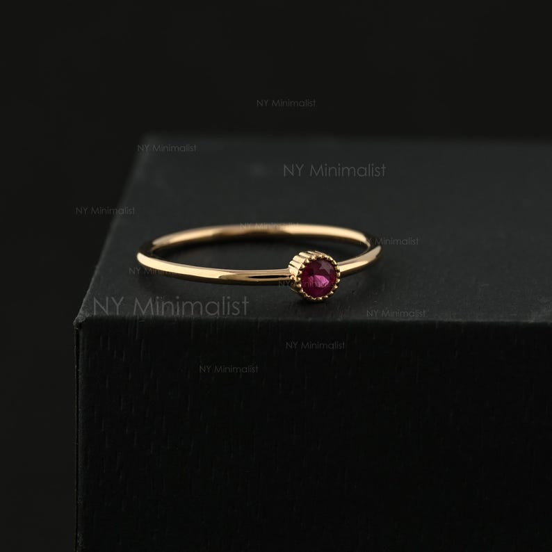 May include: A thin gold ring featuring a single, small, round, faceted pink gemstone set in a beaded bezel. The band is smooth and polished, showcasing a delicate design with a vibrant, circular pink stone as the central focal point.
