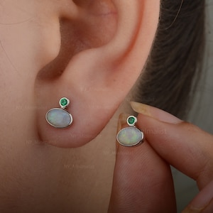 Op de afbeelding: Een paar zilveren oorbellen met ovale witte opaalstenen en kleine groene edelstenen. De oorbellen worden getoond op het oor van een persoon.
