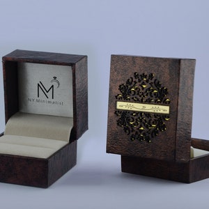 May include: A dark brown textured ring box with a cream-colored interior. The open lid displays the logo NY Minimalist with a ring icon. The closed lid features a decorative laser-cut circular pattern and a horizontal gold-colored metal plate.