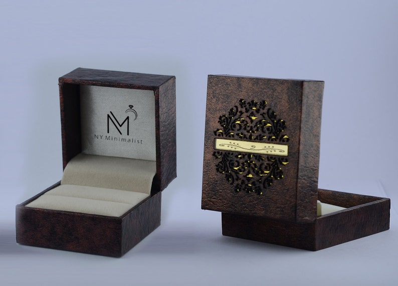 May include: A dark brown textured ring box with a cream-colored interior. The open lid displays the logo NY Minimalist with a ring icon. The closed lid features a decorative laser-cut circular pattern and a horizontal gold-colored metal plate.