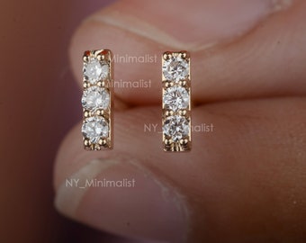 May include: A pair of gold earrings with a vertical row of three small, clear gemstones. The earrings are simple and minimalist.