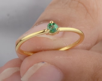 Puede incluir: Un delicado anillo de oro con una pequeña piedra preciosa redonda de color verde esmeralda engastada en el centro.