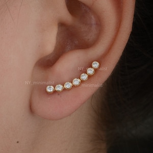 以下が含まれることがあります： 透明な丸いキュービックジルコニアの石が並んだ、金の耳飾り。耳飾りはカーブしていて、耳の上部に置かれます。