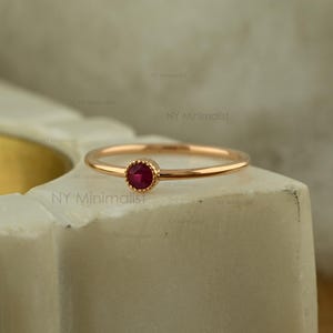 May include: A thin gold band ring featuring a single, small, round, deep red ruby gemstone set in a delicate, beaded bezel setting. The ring has a polished, smooth finish and a simple, slender design that highlights the vibrant red stone.