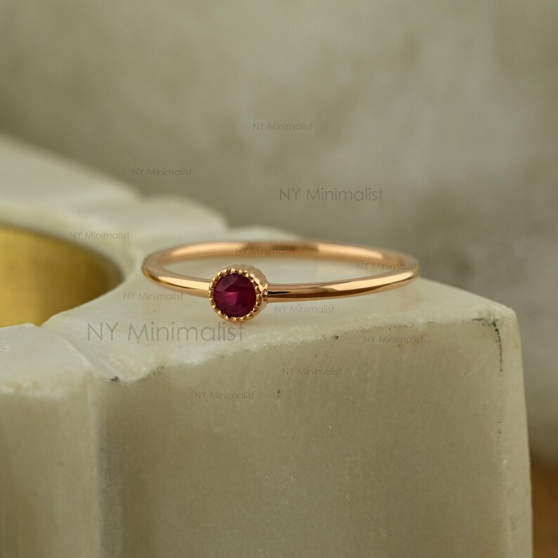 May include: A thin gold band ring featuring a single, small, round, deep red ruby gemstone set in a delicate, beaded bezel setting. The ring has a polished, smooth finish and a simple, slender design that highlights the vibrant red stone.