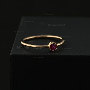May include: A thin gold ring featuring a single, small, round, deep red gemstone set in a beaded bezel setting. The band is smooth and circular, and the text NY Minimalist is visible in the background, repeating across the dark surface.