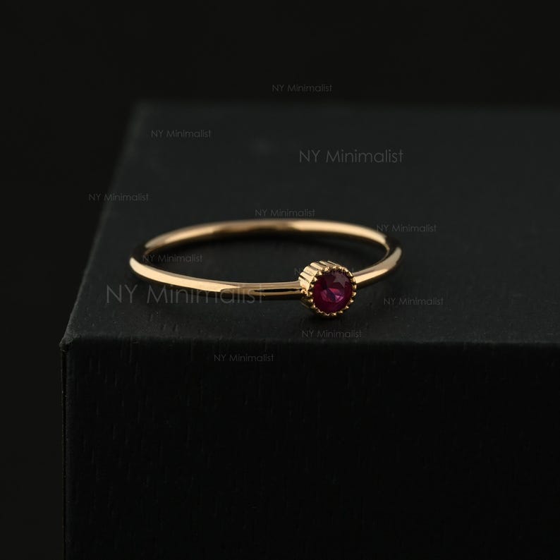 May include: A thin gold ring featuring a single, small, round, deep red gemstone set in a beaded bezel setting. The band is smooth and circular, and the text NY Minimalist is visible in the background, repeating across the dark surface.