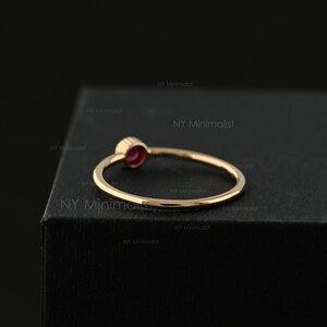 May include: A thin gold ring featuring a small, round, deep pink gemstone set in a bezel setting. The band is smooth and polished, showcasing a simple circular design. The text NY Minimalist is repeated across the dark background surface.