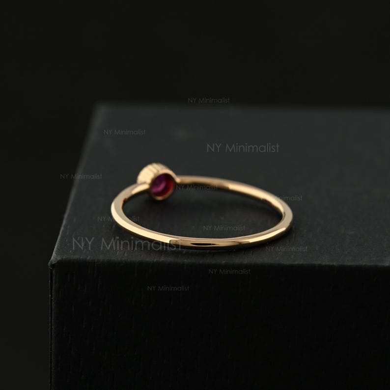 May include: A thin gold ring featuring a small, round, deep pink gemstone set in a bezel setting. The band is smooth and polished, showcasing a simple circular design. The text NY Minimalist is repeated across the dark background surface.