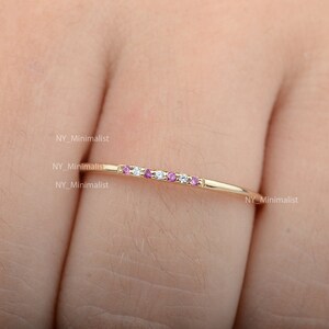 May include: A delicate gold band ring with a row of alternating pink and white gemstones. The ring is shown on a finger.
