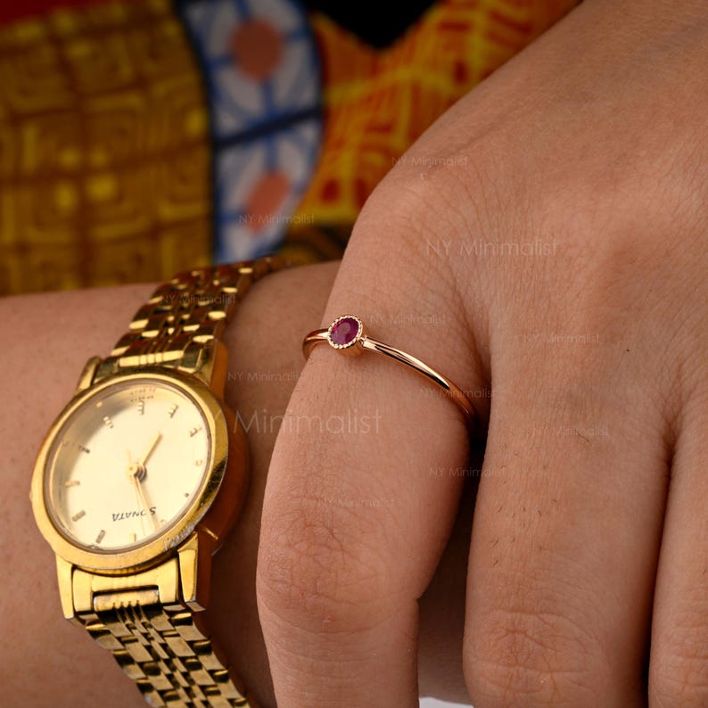 May include: A thin gold ring featuring a small, round, bezel-set red gemstone, worn on a finger next to a gold-tone analog wristwatch with a metallic link band and a round face with gold markers and hands.