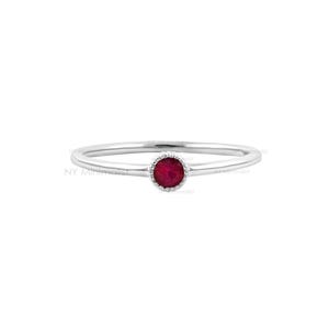 May include: A thin silver-toned ring featuring a single round, faceted, deep red ruby gemstone set in a delicate, beaded metal bezel. The band is smooth and slender, centered around the gemstone. The text NY Minimalist is visible in the background.