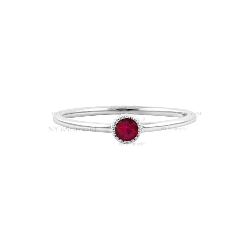 May include: A thin silver-toned ring featuring a single round, faceted, deep red ruby gemstone set in a delicate, beaded metal bezel. The band is smooth and slender, centered around the gemstone. The text NY Minimalist is visible in the background.