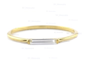 May include: A delicate gold ring featuring a slender band and a rectangular, clear gemstone. The gemstone is set horizontally in the center of the ring. The ring has a minimalist design.
