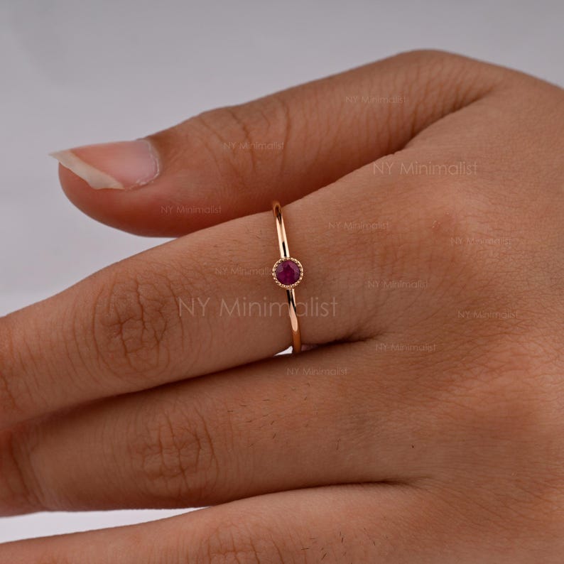 May include: A thin gold ring featuring a single, small, round, deep pink ruby gemstone set in a bezel setting with a delicate beaded edge. The band is smooth and slender, showcasing a minimalist design that highlights the vibrant gemstone.