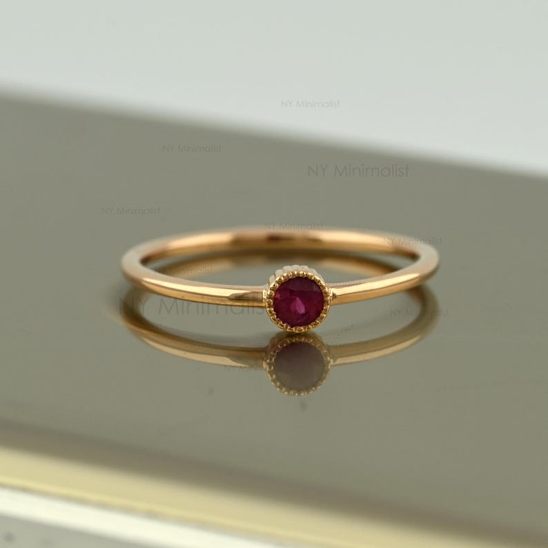 May include: A thin gold ring featuring a single round, faceted, deep pink ruby gemstone set in a beaded bezel setting. The band is smooth and polished, showcasing a delicate design with the text NY Minimalist visible in the background.