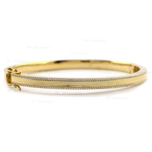 Puede incluir: Una pulsera dorada con una sección central rectangular y una fila de pequeñas piedras transparentes a cada lado. La pulsera tiene un cierre de broche y está sobre un fondo blanco.