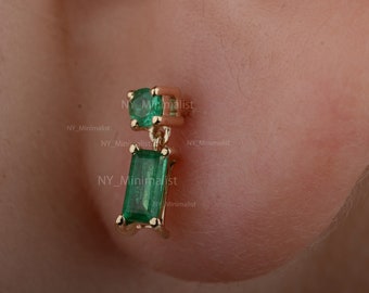 May include: Gold drop earrings with emerald green gemstones. The top stone is round, the lower one rectangular. The earrings are shown against a blurred skin background. The earrings are a stylish accessory.
