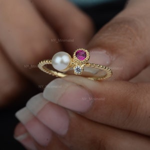 Op de afbeelding: Een gouden ring met een roze steen, een parel en een kleine heldere steen. De ring wordt gedragen aan een vinger.