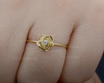 Può includere: Un delicato anello d'oro con un design a forma di rosa. La rosa presenta petali dettagliati e un piccolo diamante scintillante al centro. L'anello è mostrato su un dito, evidenziando il suo stile elegante e femminile.