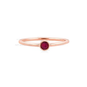 May include: A thin rose gold ring featuring a single, small, round, deep red ruby gemstone set in a delicate, beaded bezel setting. The band is smooth and polished, showcasing a minimalist design with the text NY Minimalist visible.