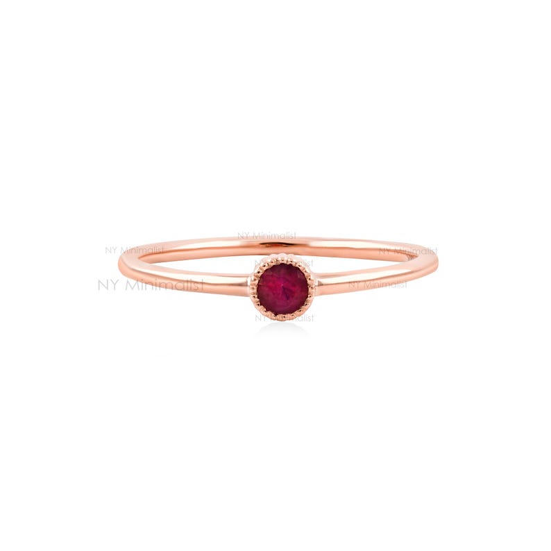 May include: A thin rose gold ring featuring a single, small, round, deep red ruby gemstone set in a delicate, beaded bezel setting. The band is smooth and polished, showcasing a minimalist design with the text NY Minimalist visible.