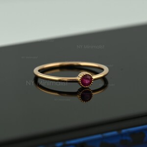 May include: A thin gold ring featuring a single, small, round, faceted red ruby gemstone set in a beaded bezel setting. The ring has a smooth, polished gold band and is positioned to show its reflection on a dark, reflective surface.