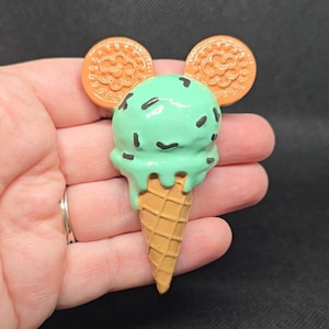 May include: A mint green ice cream cone with chocolate sprinkles and two orange cookie ears. The ice cream is dripping down the waffle cone. The design is held in a hand.