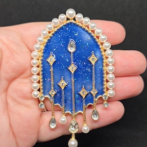 May include: A blue and gold brooch with a celestial design. The brooch features a blue background with glitter, gold accents, and faux pearls. The design includes diamond-shaped and teardrop-shaped embellishments.