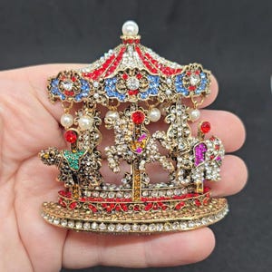 May include: A detailed carousel brooch with red, white, blue, and gold accents. The miniature carousel features three jeweled horses and a decorative canopy, adorned with faux pearls and sparkling crystals. A whimsical accessory.