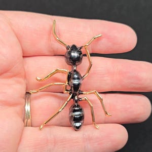 May include: A detailed, metallic ant figurine with a black body and gold-colored legs and antennae. The ant is posed as if walking, with its legs spread out. The figurine is held in a hand, showcasing its size and detail.