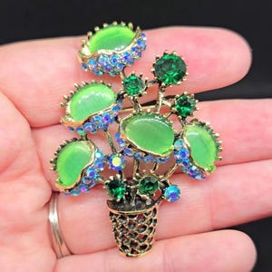 May include: A gold-tone metal brooch shaped like a potted plant, featuring five smooth, light green cabochon stones and several smaller, faceted dark green crystals. The base is a textured, open-work gold-tone pot with iridescent blue accents.