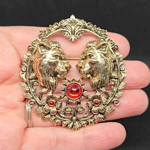 May include: A vintage gold-toned brooch featuring two lion heads facing each other. The brooch has a central red jewel, surrounded by a sunburst design, and floral accents. The brooch is oval-shaped.