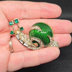May include: A gold-tone snail brooch featuring a vibrant green enamel shell with striped detailing. The body is encrusted with clear baguette and round rhinestones, while the antennae are topped with small, square and round green crystals.