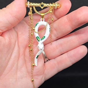 May include: A gold-toned brooch featuring a horizontal bar with a central black stone and crystal accents. A white snake pendant with green spots hangs from the bar, suspended by multiple gold chains adorned with small gold beads.
