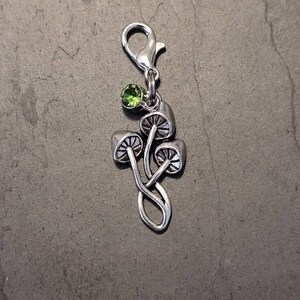 May include: A silver charm depicting three stylised mushrooms with detailed caps. A small, round, green gemstone is set above the mushrooms. The charm has a lobster clasp for attachment to a bracelet or necklace.
