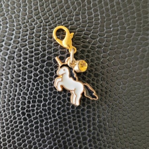 May include: A gold charm featuring a white unicorn and a yellow rhinestone. The charm is attached to a gold split ring.