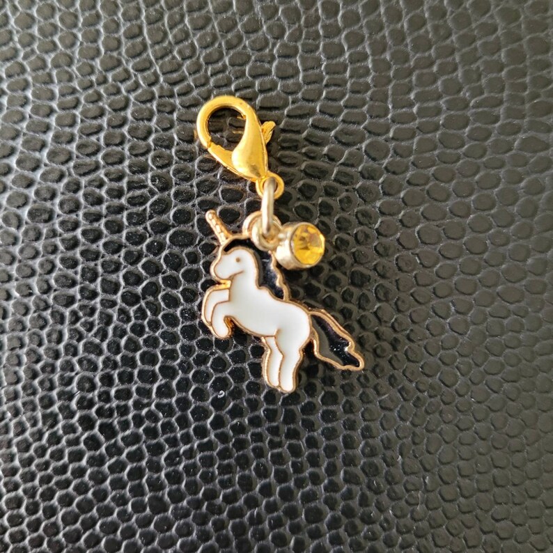 May include: A gold charm featuring a white unicorn and a yellow rhinestone. The charm is attached to a gold split ring.
