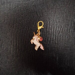 May include: A gold-tone charm featuring a white unicorn with pink accents and a pink gem. The charm has a gold-tone clip on the top.