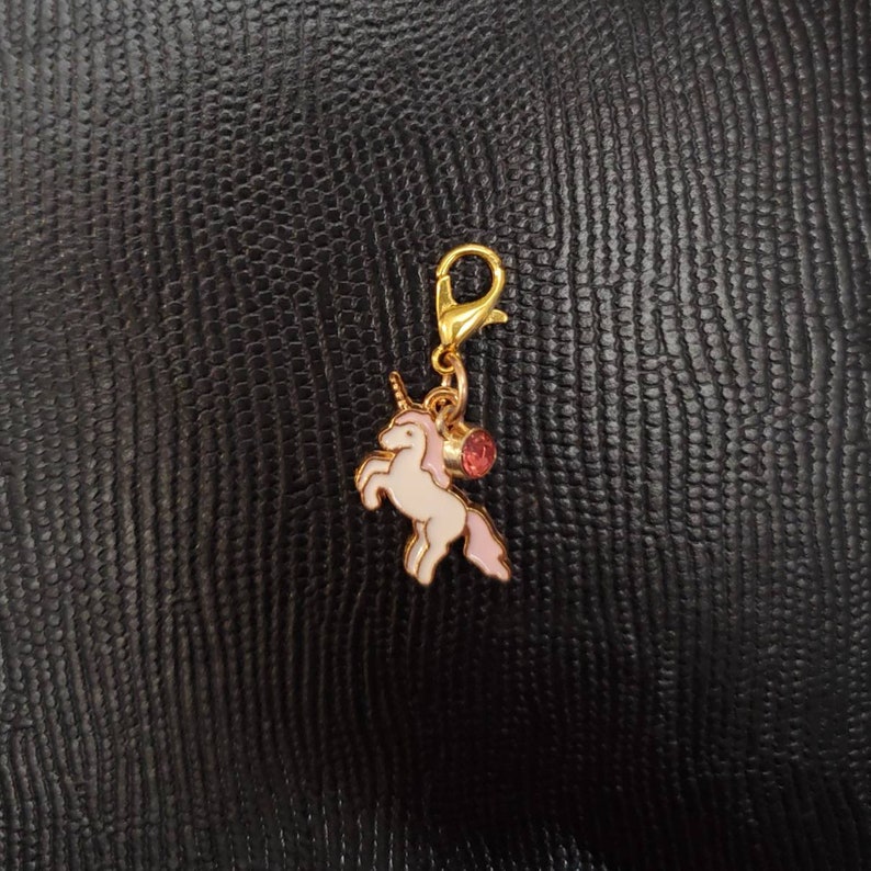 May include: A gold-tone charm featuring a white unicorn with pink accents and a pink gem. The charm has a gold-tone clip on the top.